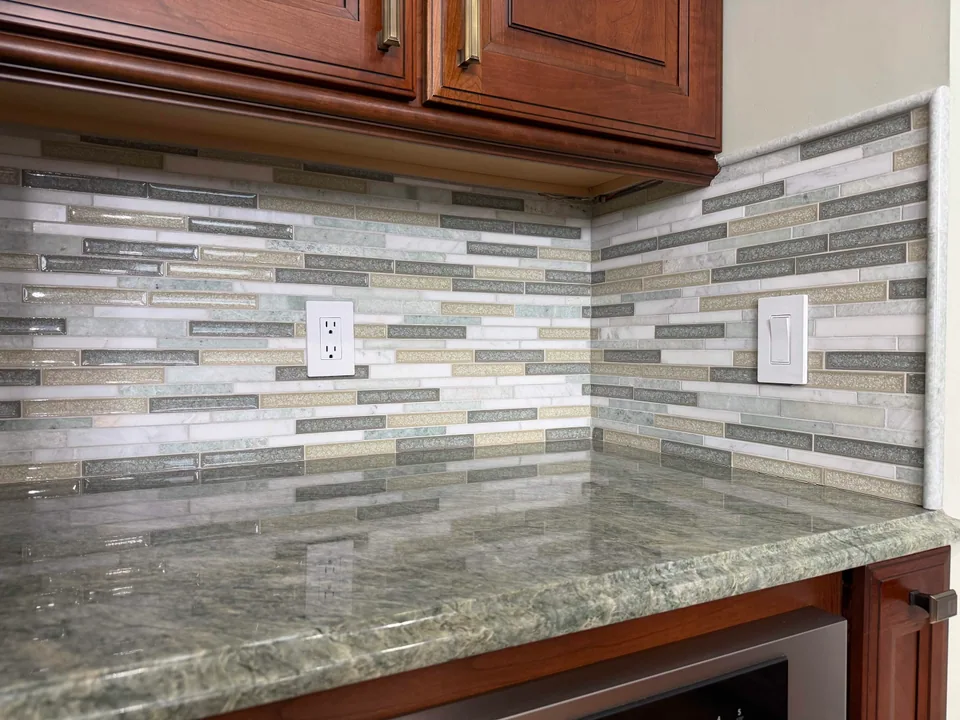 Linear backsplash tile continuing into an inside corner, with an outlet and a vertical trim at the exposed edge.