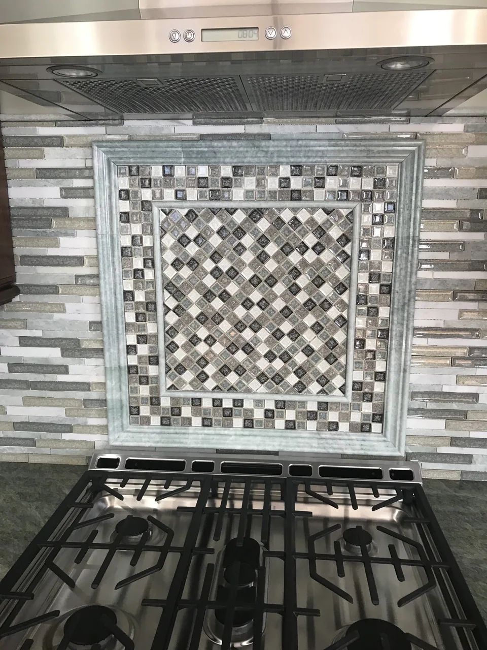 Framed kitchen backsplash panel with diagonal small tiles, a square mosaic border, and layered trim above a range.