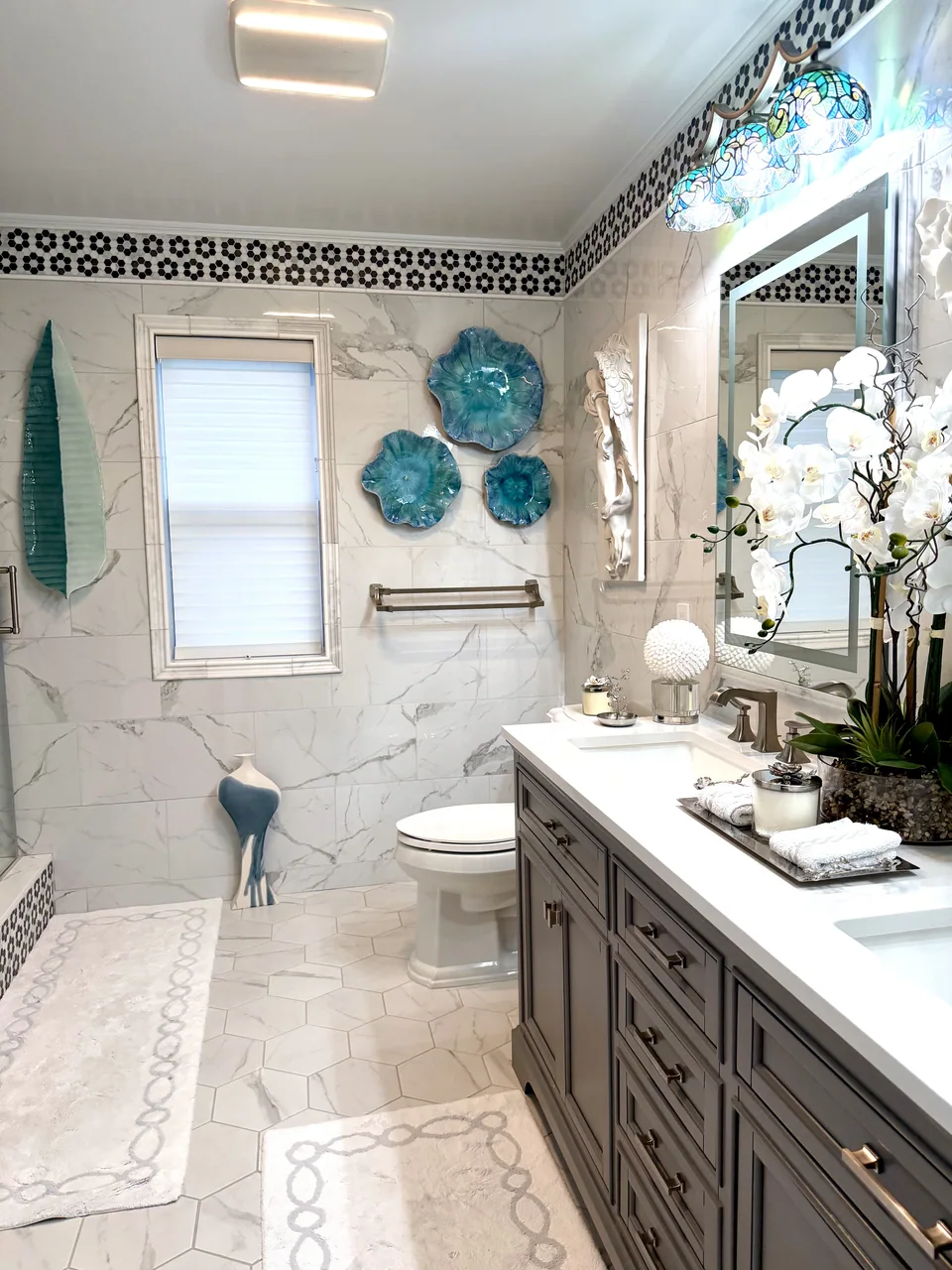 Bathroom with veined marble wall tile, a black and white mosaic border, hexagonal floor tile, a gray double vanity, and a framed window.