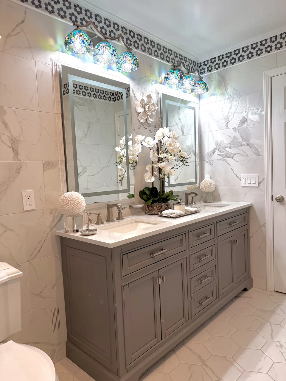 Another view of the marble bathroom showing a gray double vanity, two mirrors, decorative lights, and veined marble wall tile with a black and white mosaic border.