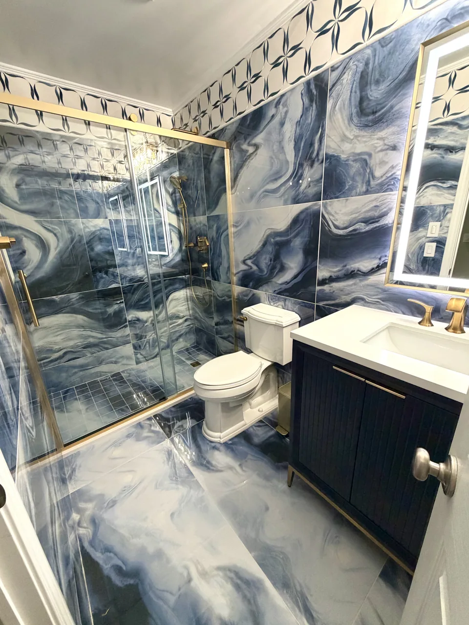 Blue and white swirling tile across bathroom walls and floor, with a glass shower enclosure, dark vanity, and gold-colored fittings.
