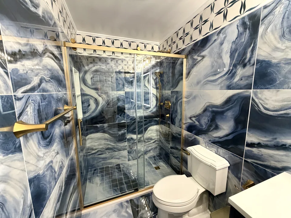 After: bathroom with blue swirling wall tile, a cream and blue patterned band, and a glass shower enclosure with gold-colored framing.
