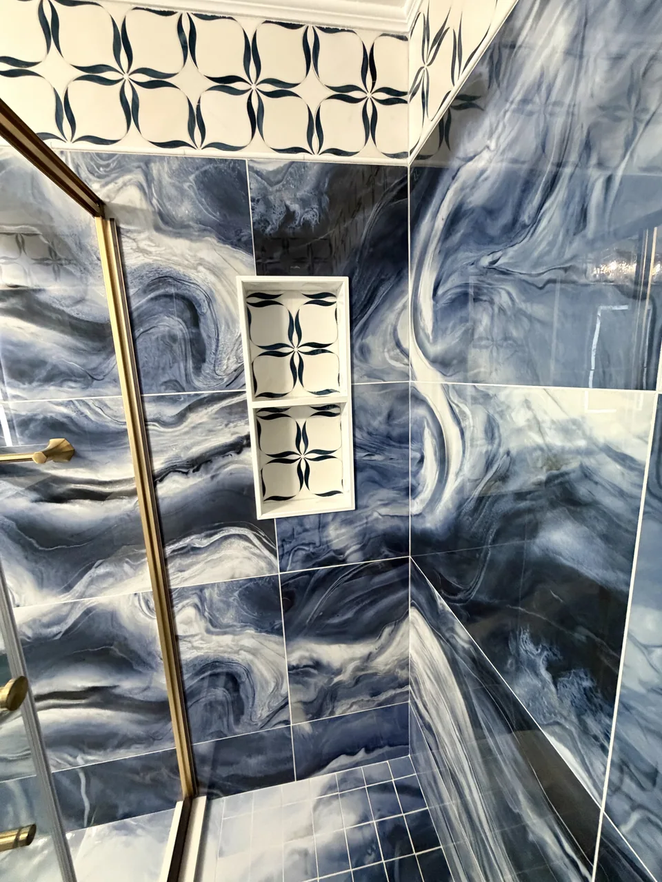 Tall shower niche with a cream and dark blue patterned backing, light border, and dividing shelf within blue swirling wall tile.