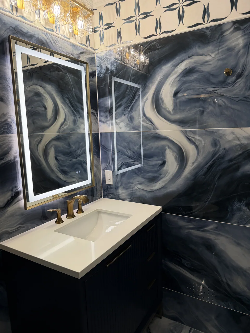 Blue swirling wall tile around a lit mirror and dark vanity, with a patterned band near the ceiling.