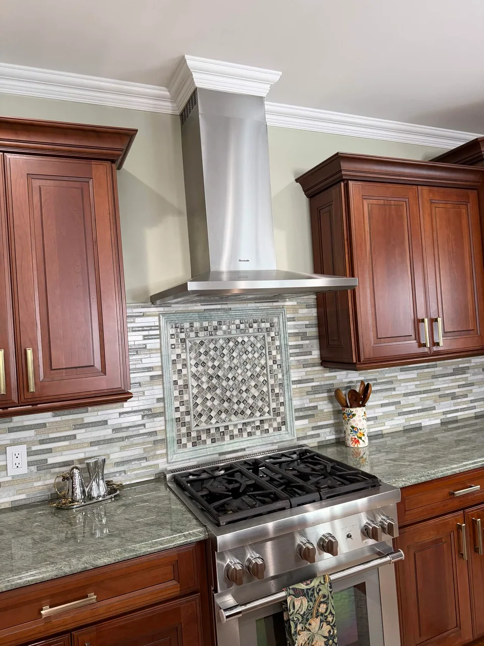 Framed patterned backsplash panel above a range, surrounded by linear tile and wood cabinets.