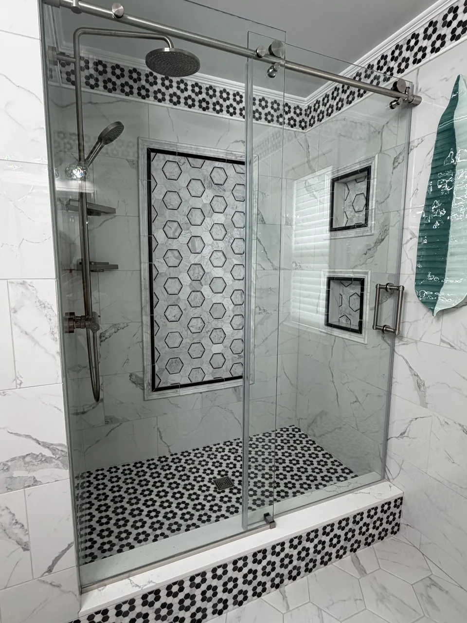 Glass-enclosed shower with light veined tile, a framed hexagonal feature panel, two recessed niches, and a black and white mosaic floor.